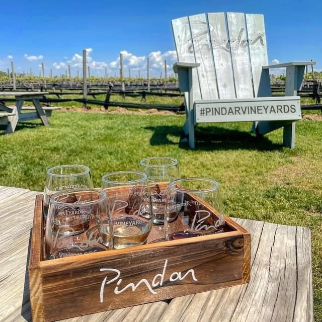 Pindar Vineyards outdoor scene with sign and wine glasses on a wooden table.