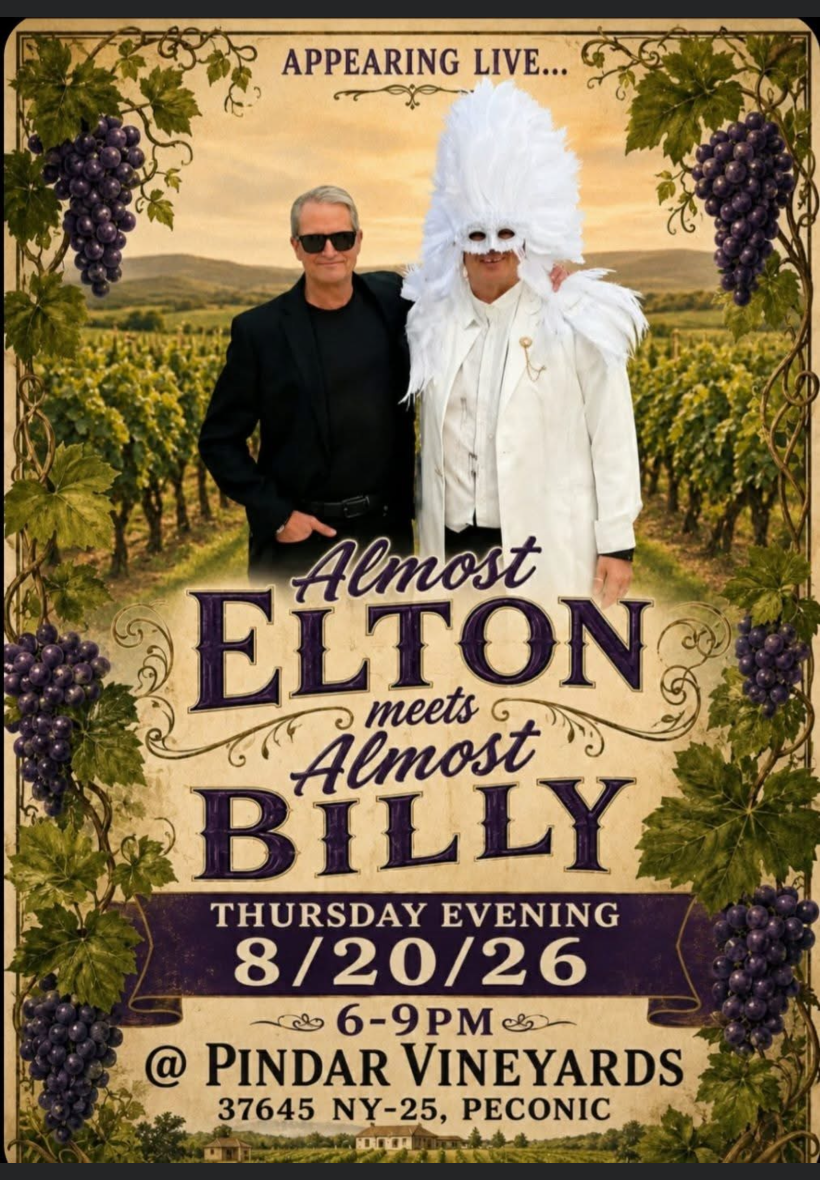 A man in black suit and sunglasses standing next to a person in a white costume with a large feathered headdress at Pindar Vi.