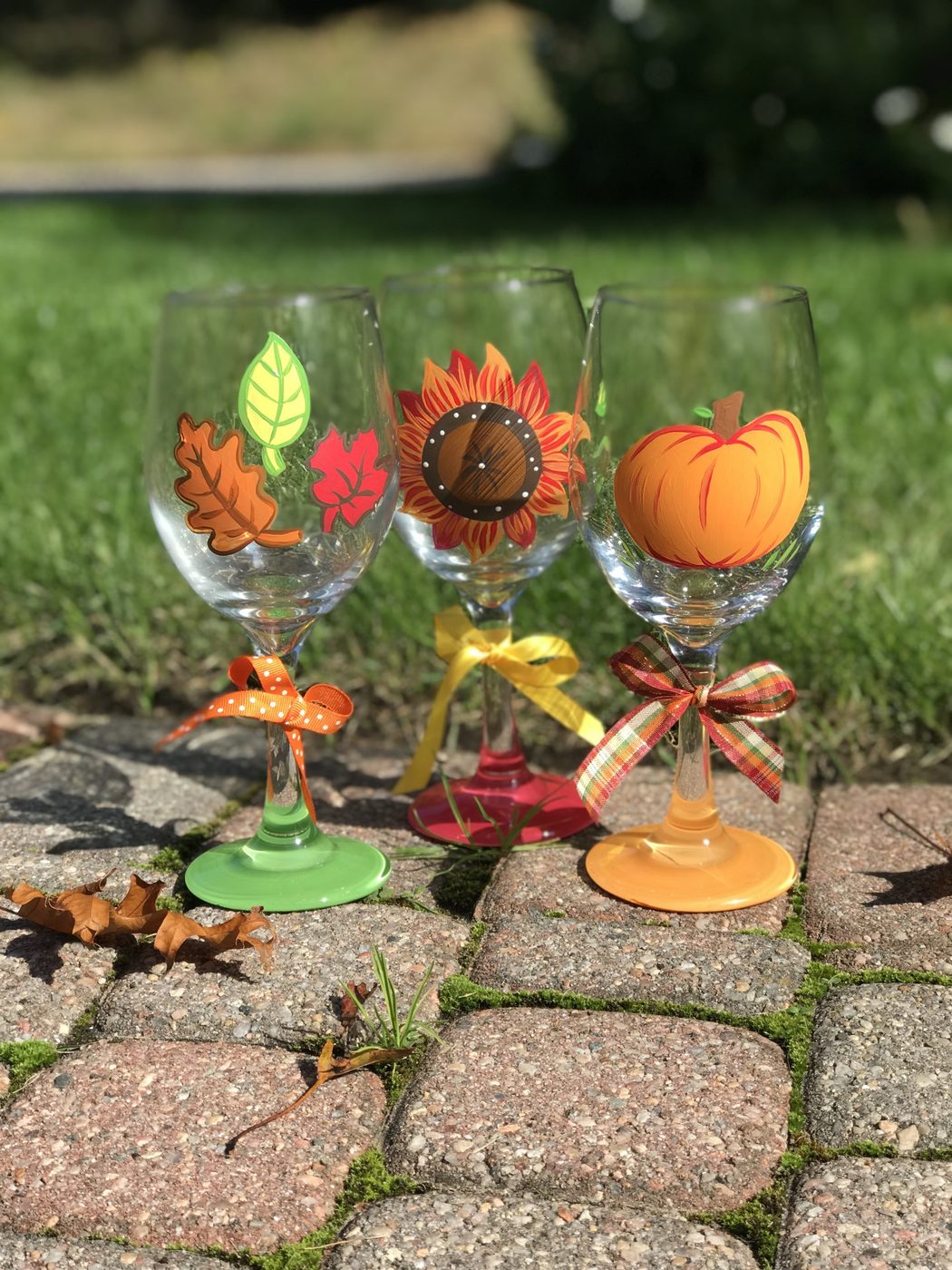 Decorative autumn-themed wine glasses with leaf, sunflower, and pumpkin designs on a brick path.